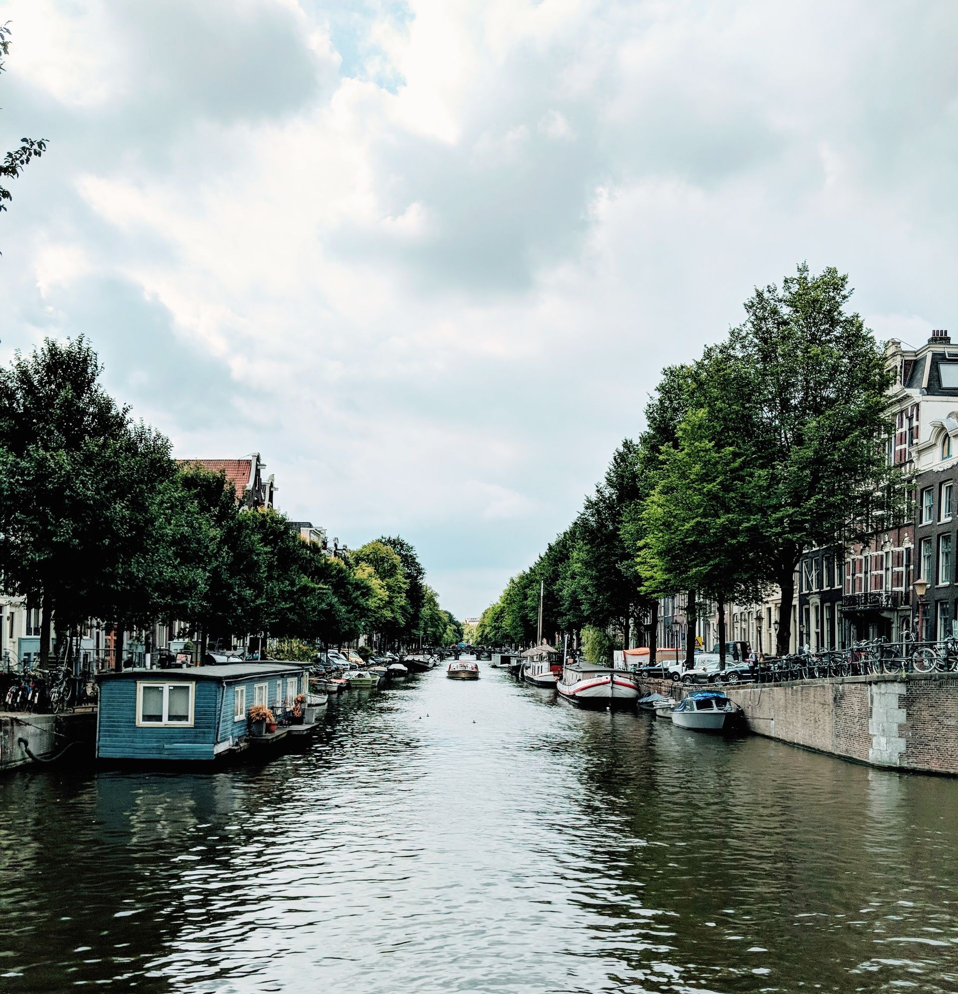 Kanal in Amsterdam, gesäumt von Bäumen, Gebäuden und Booten unter einem bewölkten Himmel.