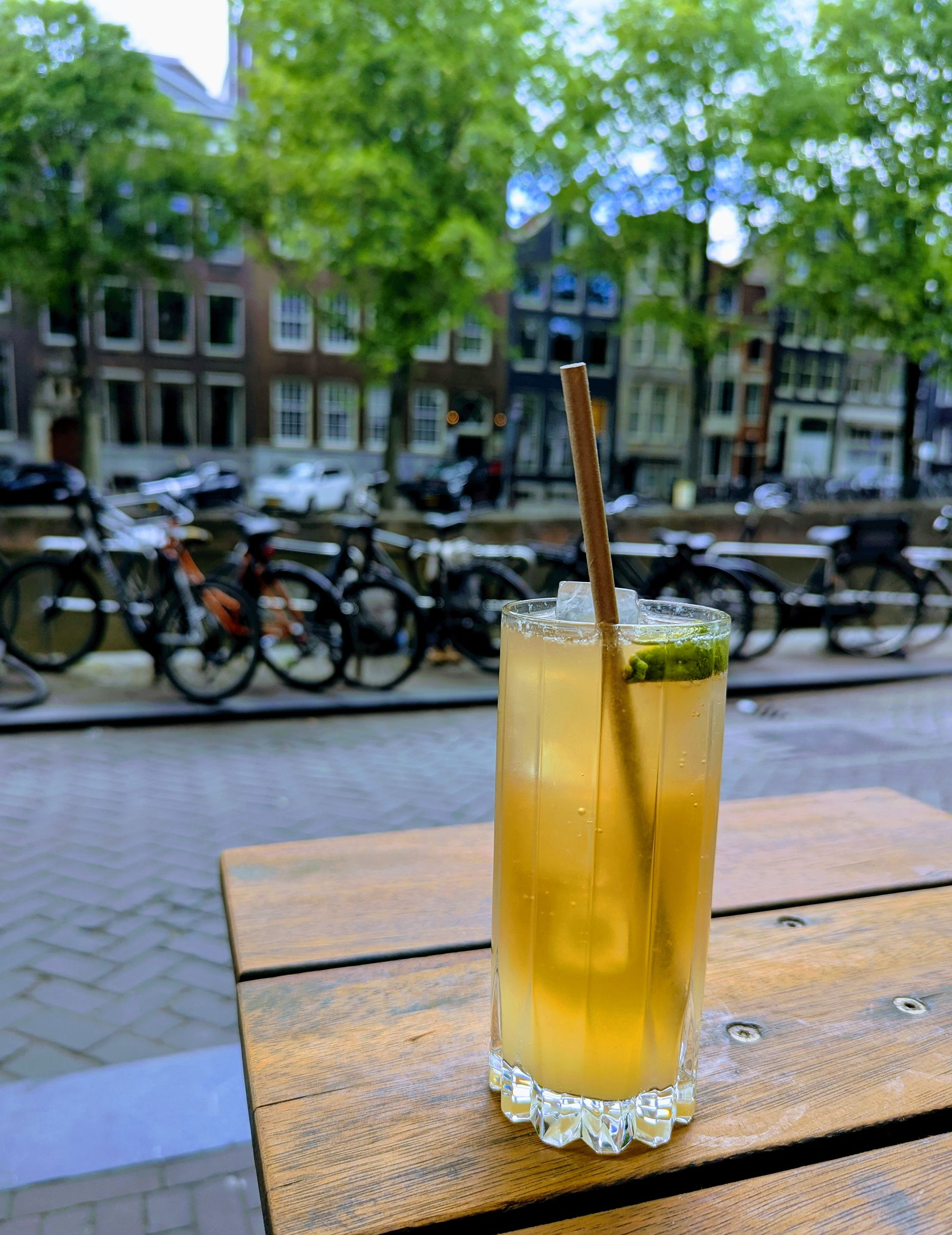 Mocktail mit Strohhalm auf einem Holztisch, Amsterdamer Straßenszene im Hintergrund: Fahrräder, Gebäude und Bäume.