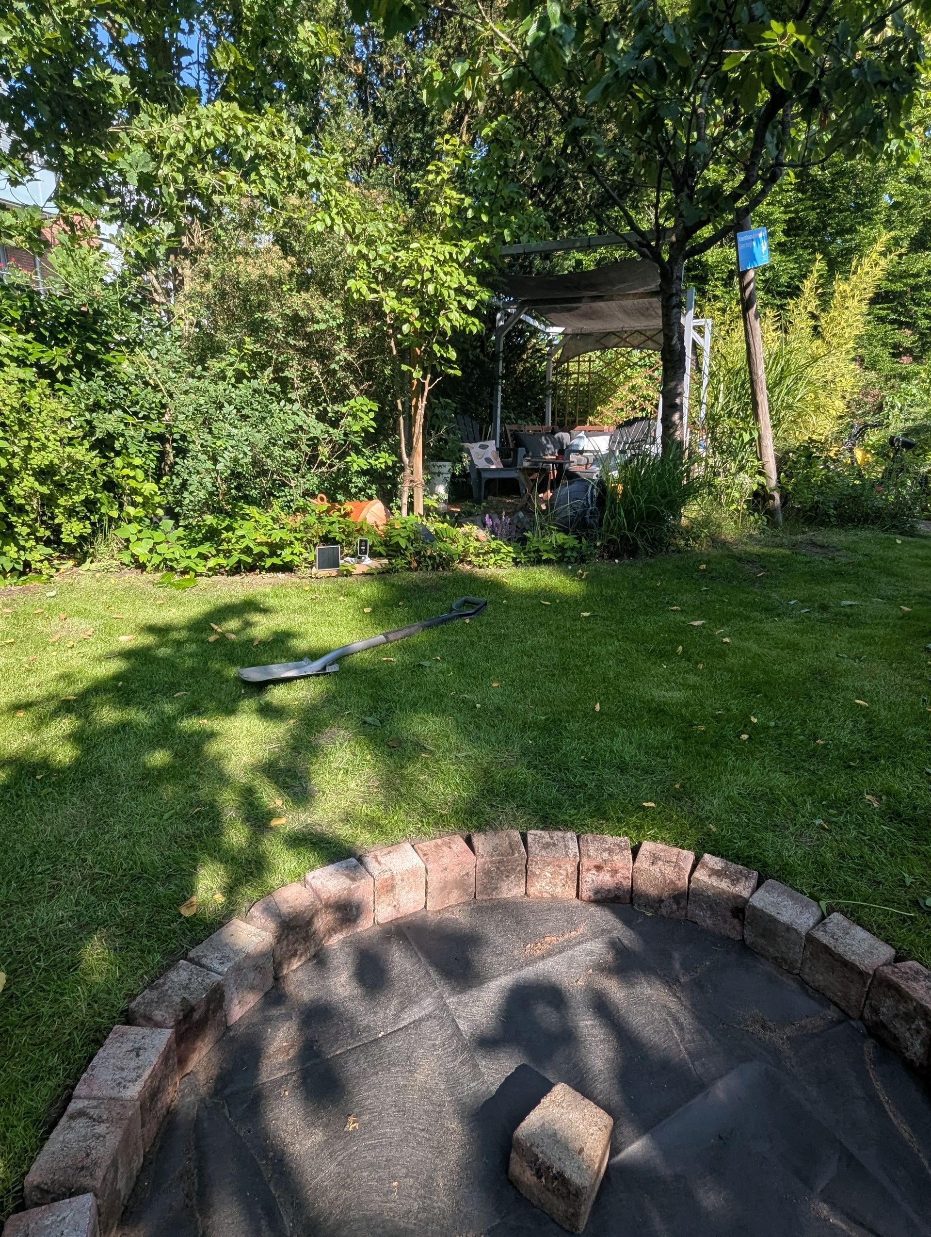 A grassy backyard with a brick-lined area in the foreground, leading to a shaded structure with foliage.