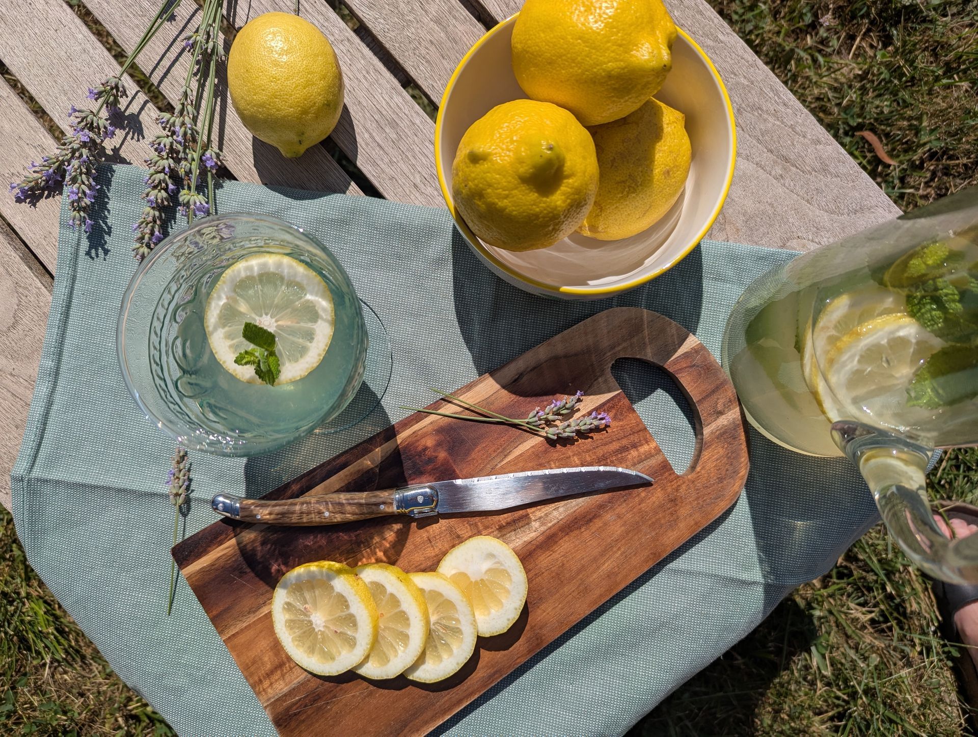 Ein Glas und eine Karaffe mit Zitronen-Lavendel-Limonade. Eine Schale mit Zitonen. EIn Holzbrett mit Zitronenscheiben und einem Messer. 