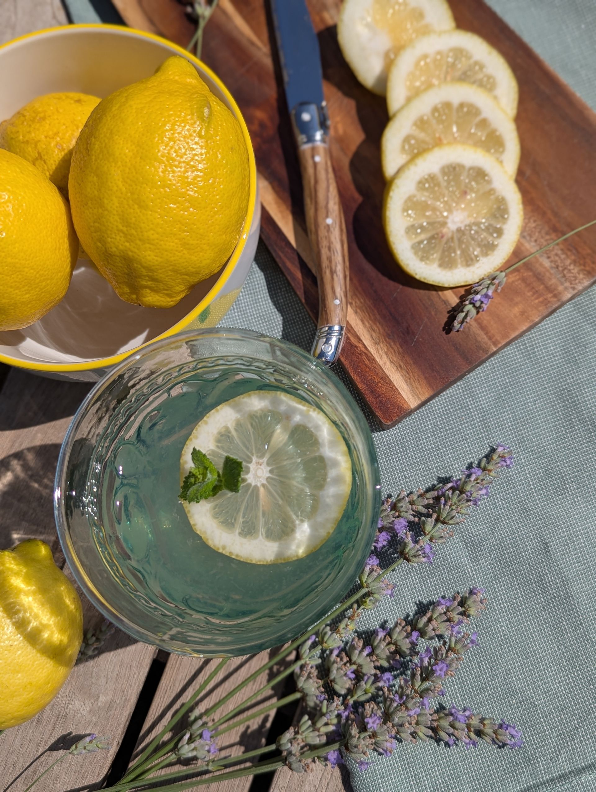 Draufsicht auf ein Glas Lavendel-Limonade, eine Schüssel Zitronen, Lavendelblüten und einem Holzbrett mit Zitronenscheiben