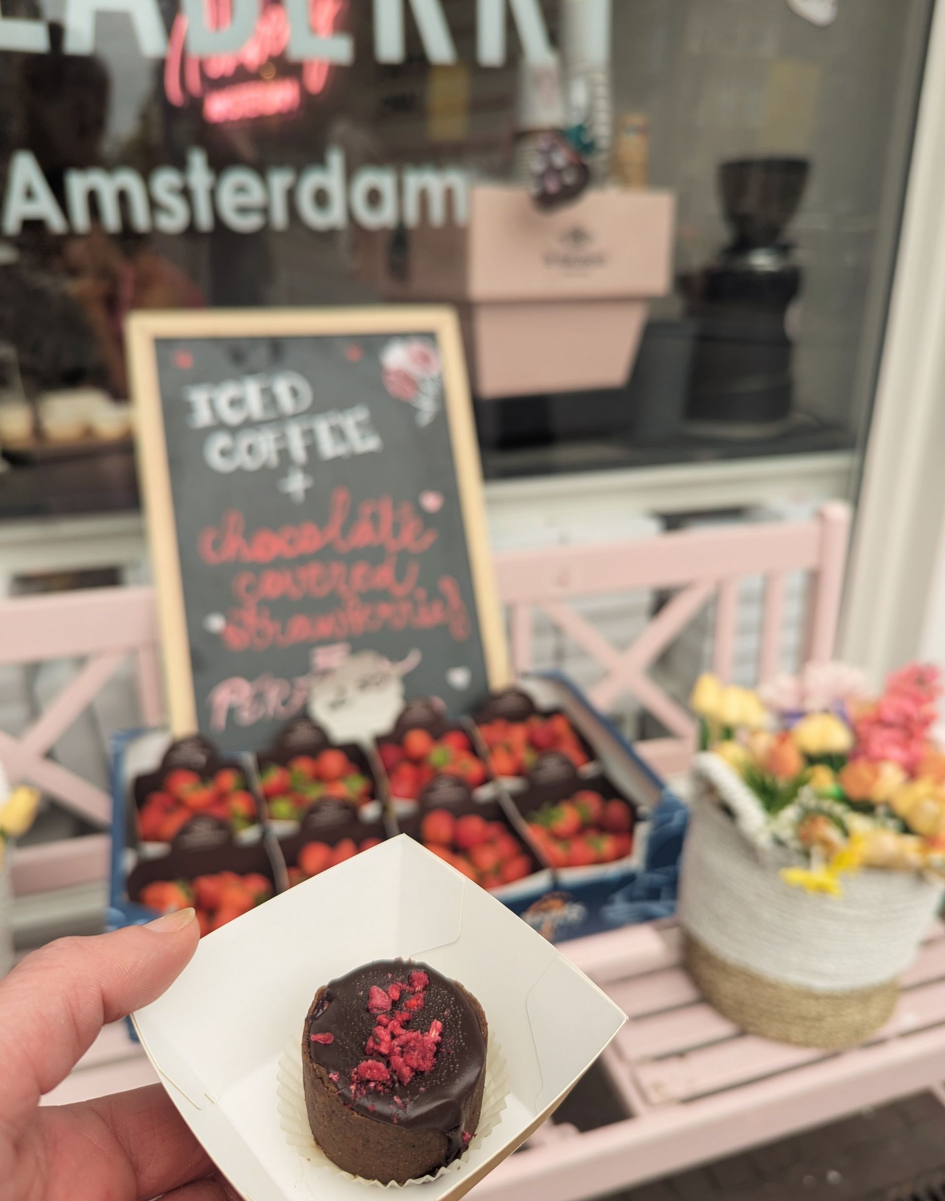 Person hält ein mit Schokolade überzogenes Erdbeerdessert. Amsterdamer Café mit Erdbeeren, Eiskaffee-Schild und rosa Akzenten im Hintergrund.