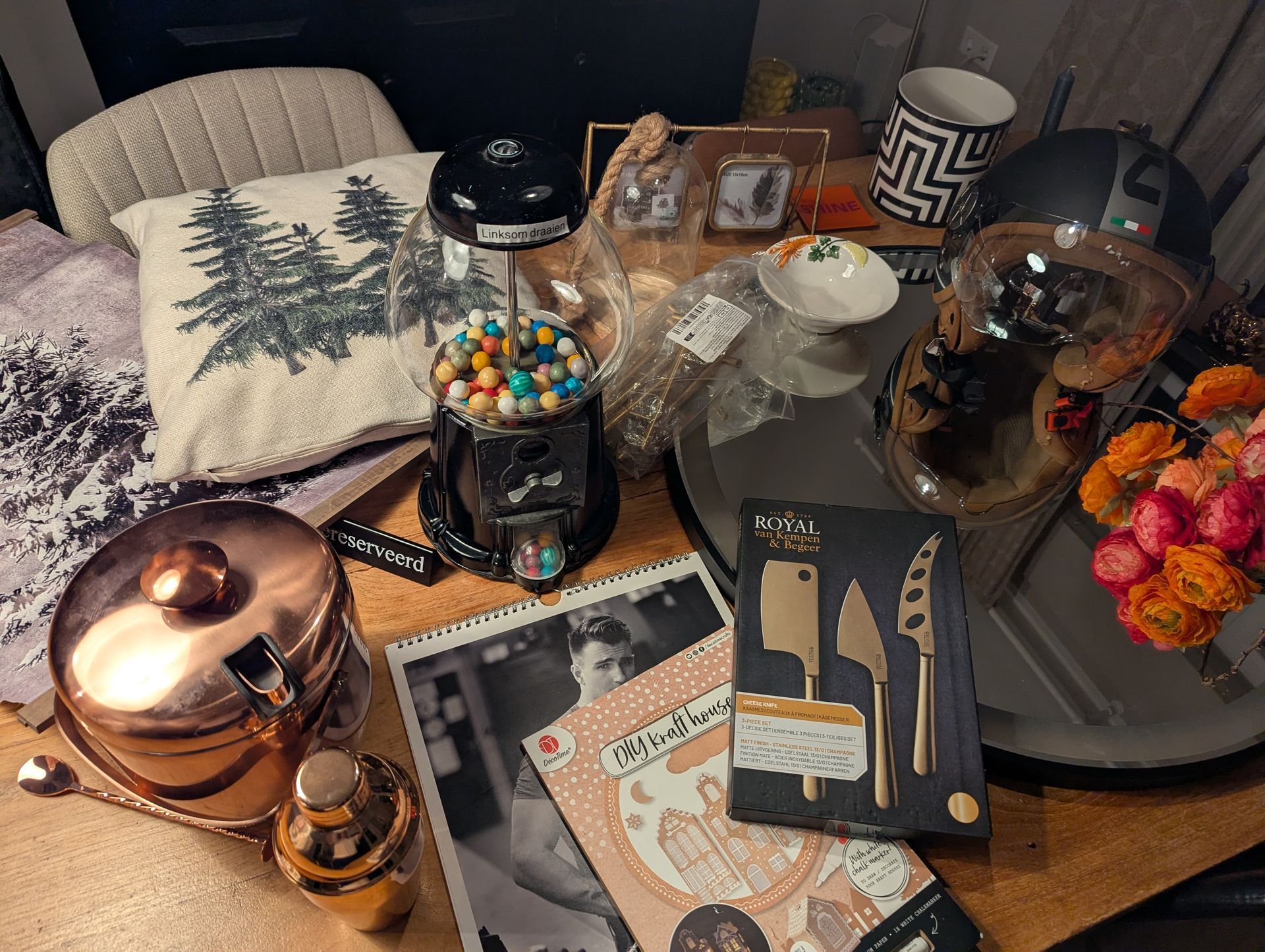 A table displays a gumball machine, copper barware, a boxed cheese knife set, a floral arrangement, and a throw pillow.