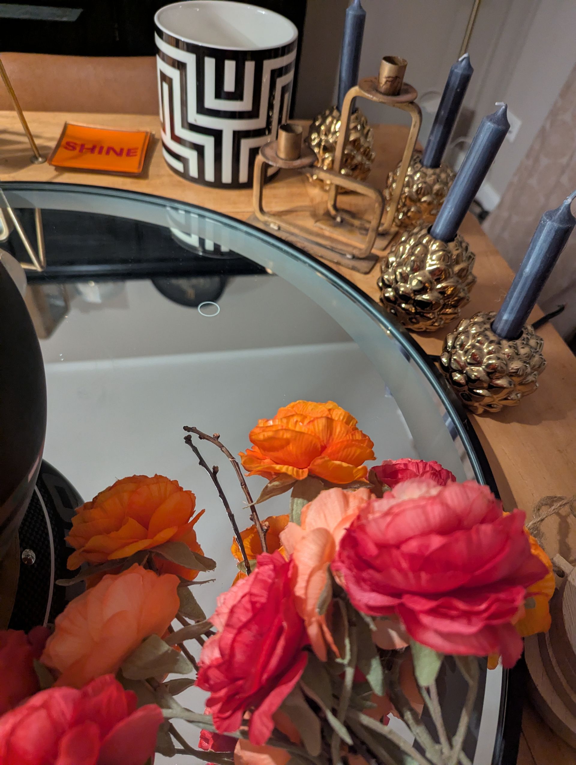 A centerpiece with artificial orange and pink flowers, a geometric vase, and gold candle holders on a round mirror tray.