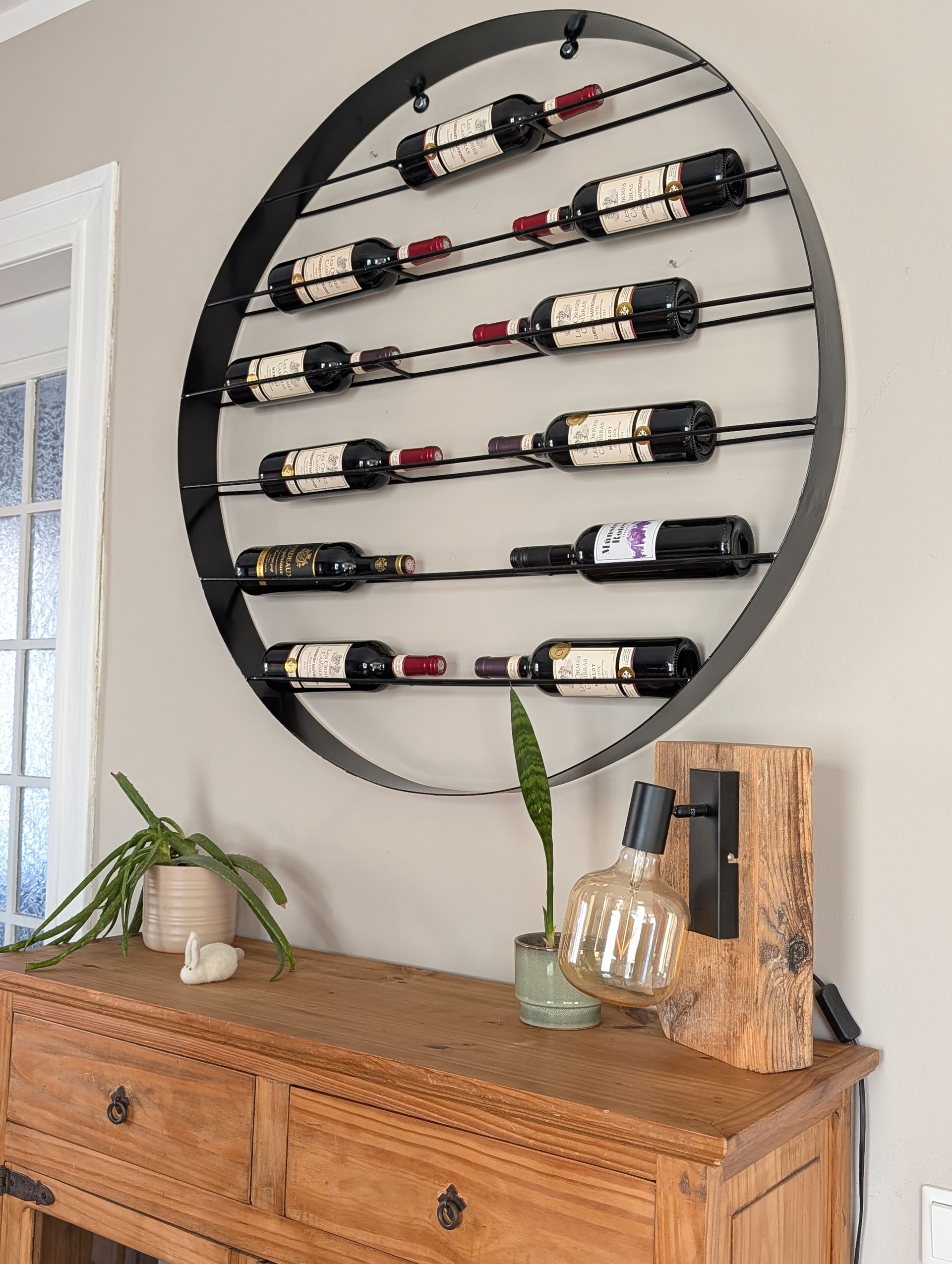 A circular black metal wall rack holding ten wine bottles, mounted above a wooden cabinet with a lamp and a plant.