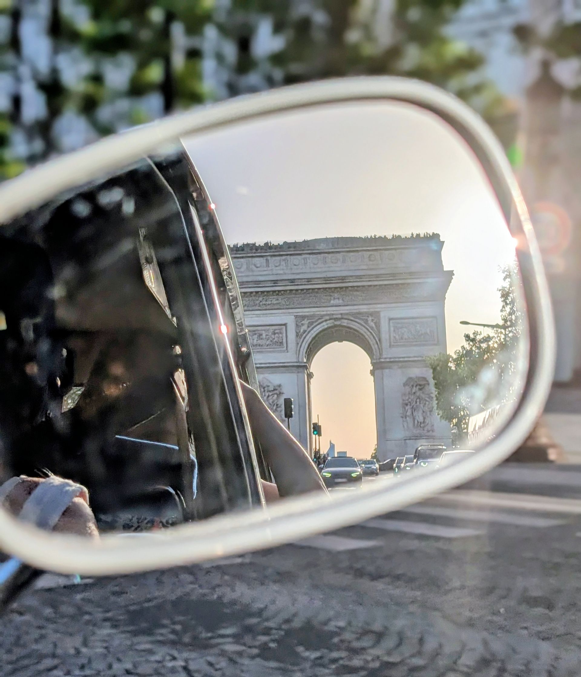 Der Arc de Triomphe spiegelt sich im Sonnenuntergang im Seitenspiegel eines Autos, dahinter ist eine Straße zu sehen.