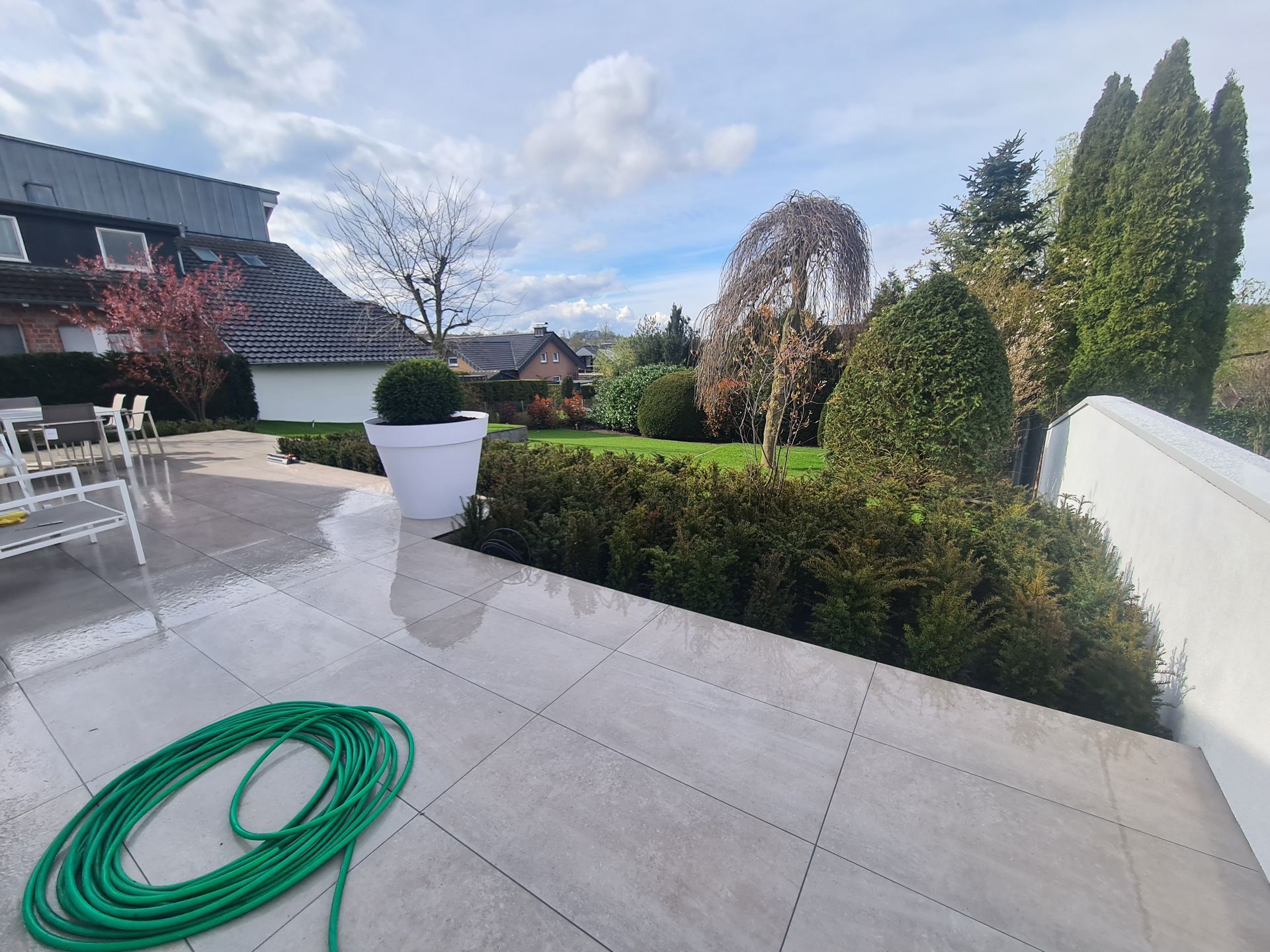Ein grüner Schlauch liegt auf einer gefliesten Terrasse neben einer Topfpflanze