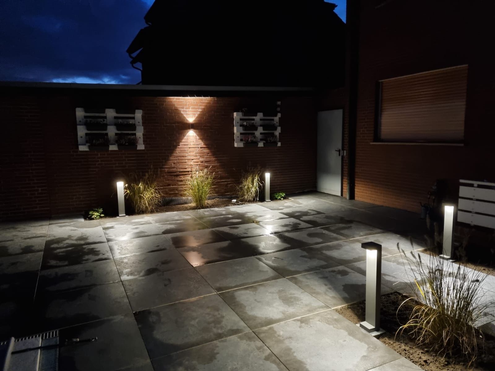Hinter einer Terrasse befindet sich eine Backsteinmauer, die nachts beleuchtet ist