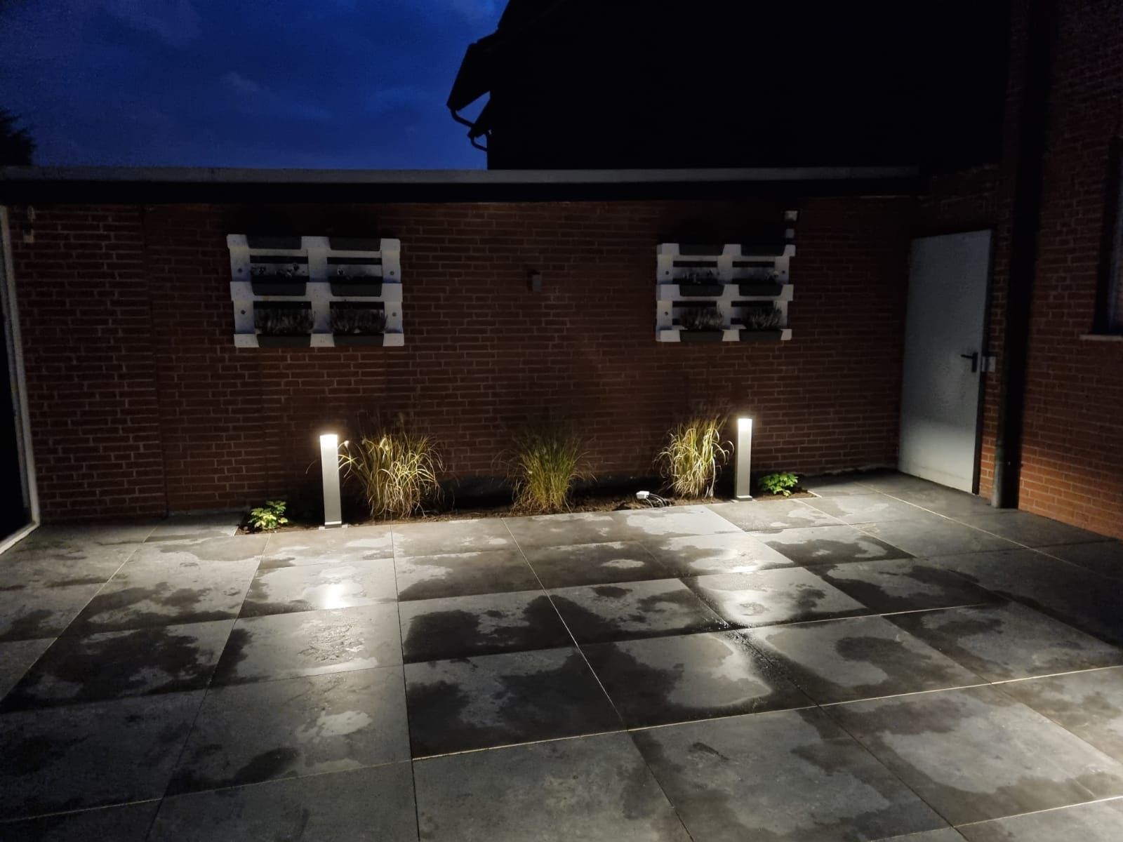 Eine Ziegelmauer ist nachts beleuchtet und davor befindet sich eine Terrasse