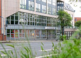 Gebäude mit Glasfassade, großen Fenstern, Bürgersteig und Bäumen davor – Praxis für Strahlentherapie