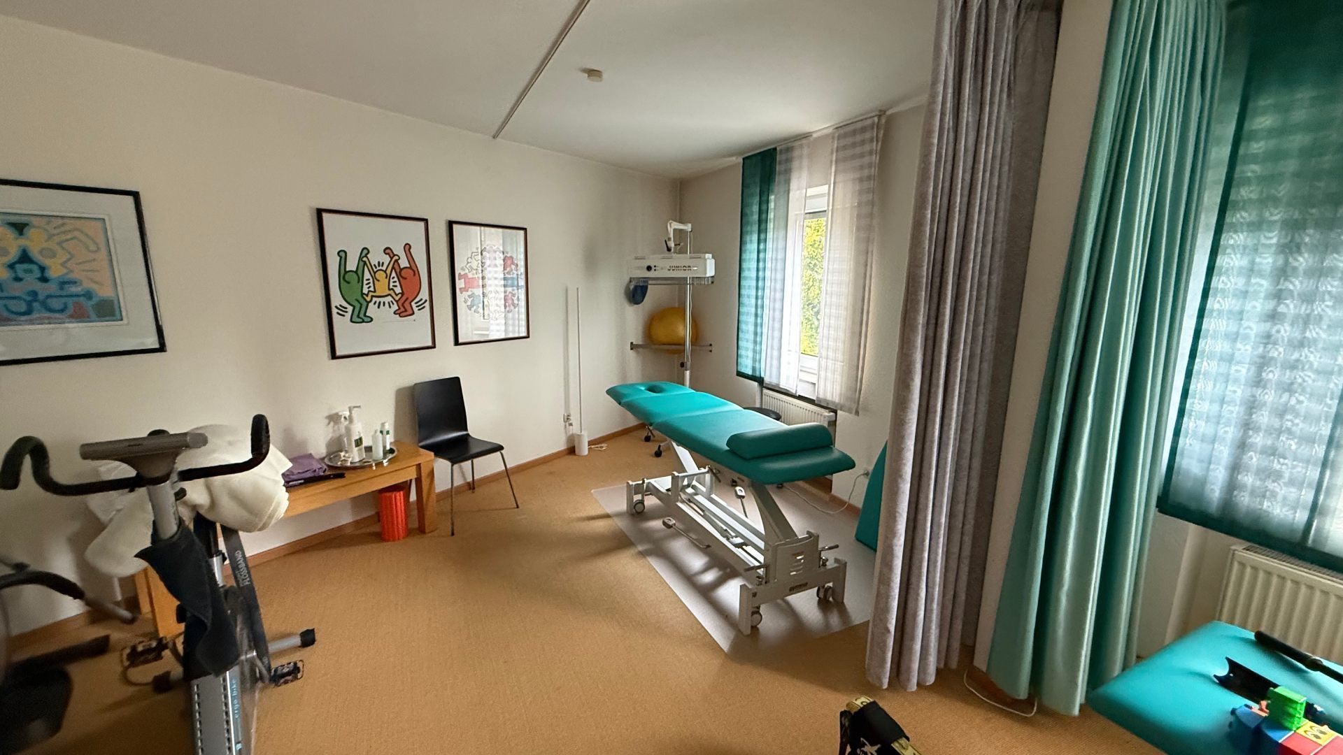 Behandlungsraum mit Therapieliege, Trainingsgeräten und großen Fenstern.