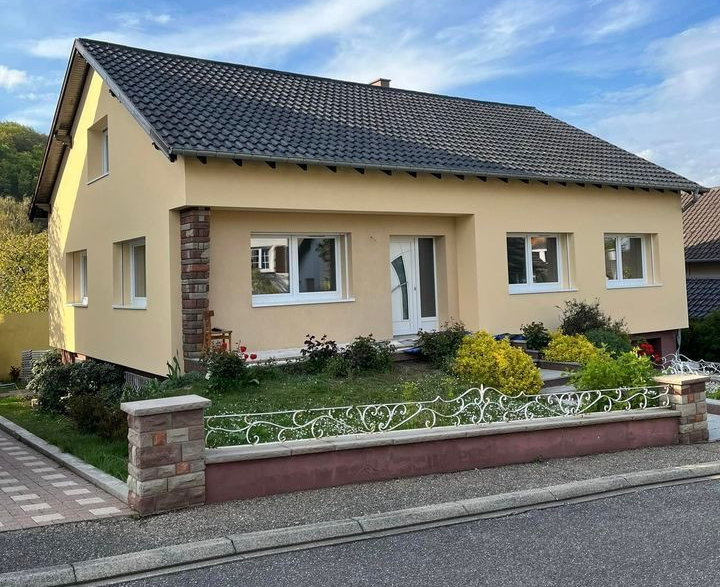 Maison beige avec toit gris foncé, fenêtres à cadre blanc et une petite cour avant avec des arbustes.