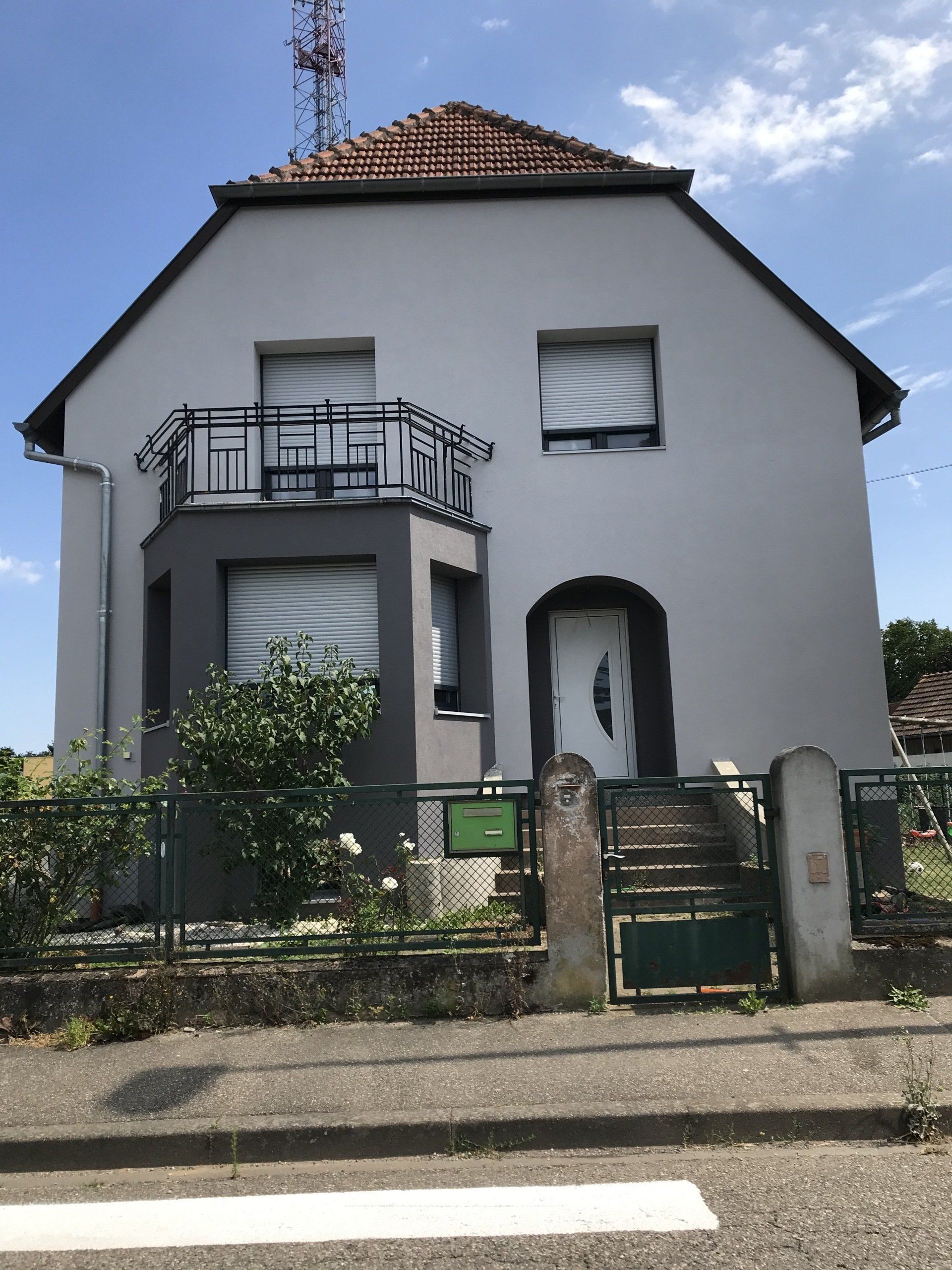 Maison à deux étages avec des accents gris, un balcon et un toit en tuiles rouges.