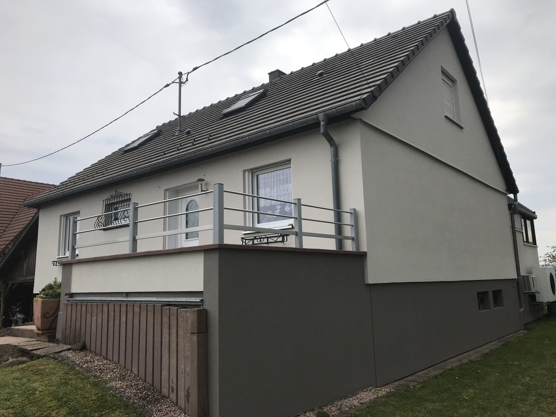 Maison au toit gris, murs blancs et partie basse grise. Balcon avec balustrade argentée, clôture en bois.