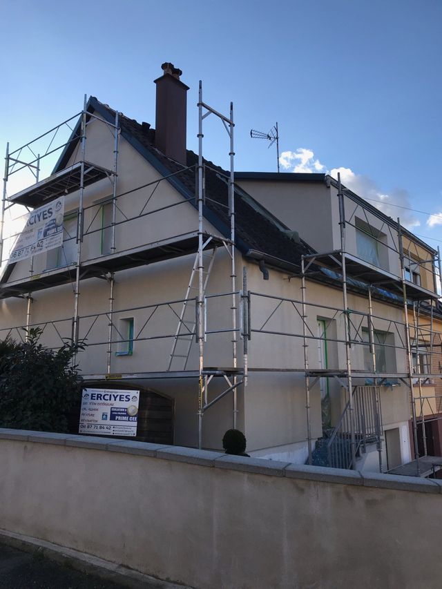 Maison avec échafaudage, façade beige clair, toit foncé et cheminée.