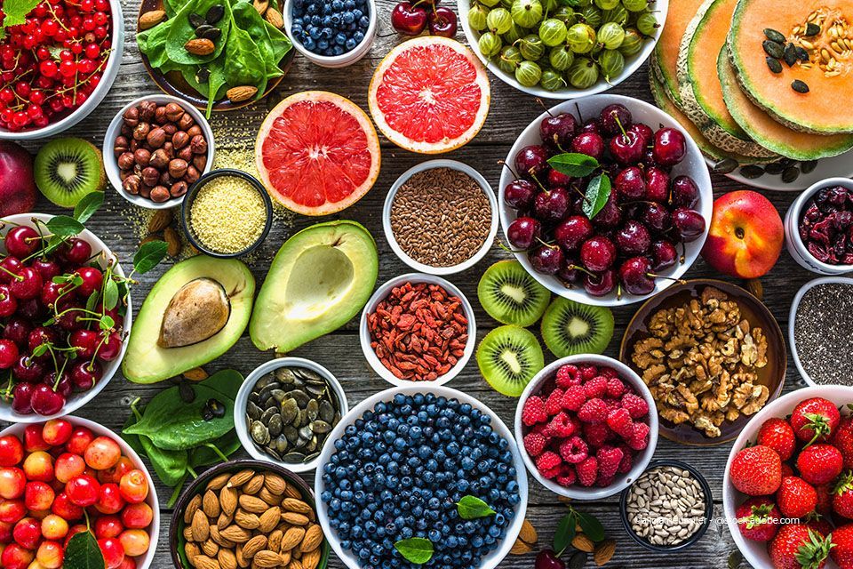 Draufsicht auf mit verschiedenen Früchten, Nüssen und Samen gefüllte Schalen auf einer Holzoberfläche, die eine bunte Auswahl gesunder Lebensmittel präsentieren.