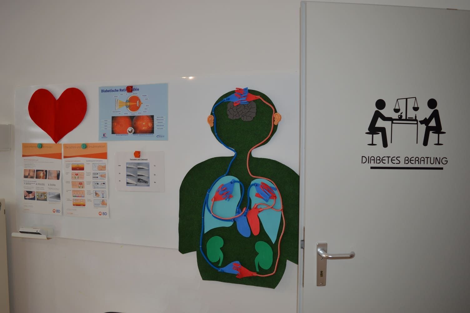 Wand mit Gesundheitsdiagrammen, Herzform und Tür mit der Silhouette einer „Diabetes-Sprechuhr“.