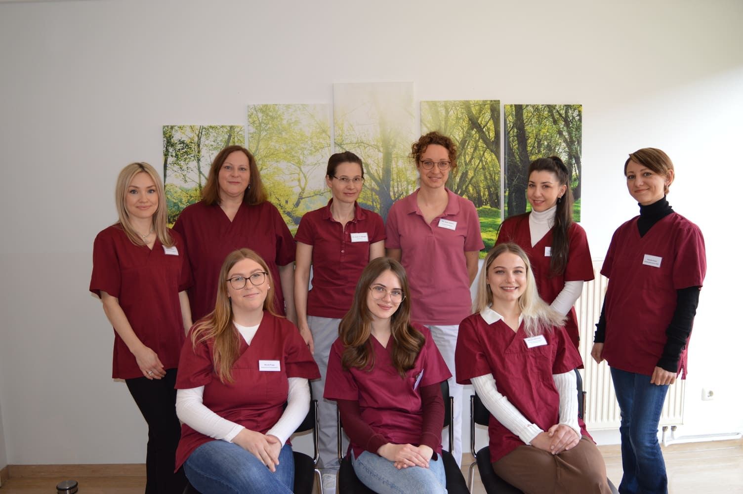 Eine Gruppe von neun Frauen in kastanienbraunen OP-Kleidern posiert vor einem Kunstwerk mit Naturmotiven.
