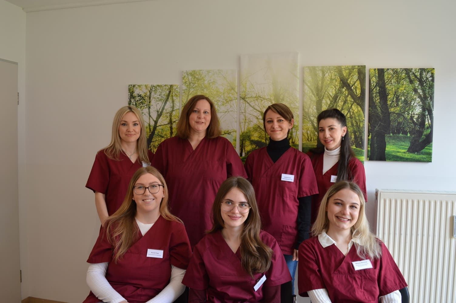 Sieben Mitarbeiter des Gesundheitswesens in kastanienbrauner OP-Kleidung posieren in einer Klinik; Kunst an der Wand.