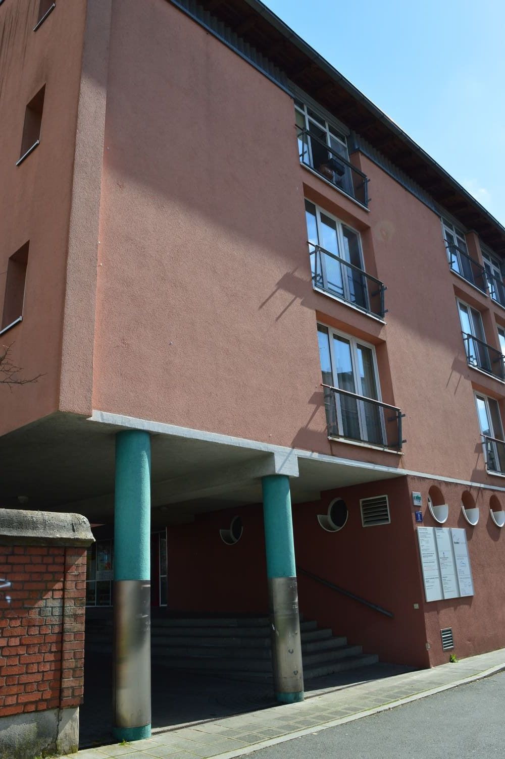 Mehrstöckiges braunes Gebäude mit blaugrünen Säulen, die ein Vordach über einem zurückgesetzten Eingang mit Treppe stützen.