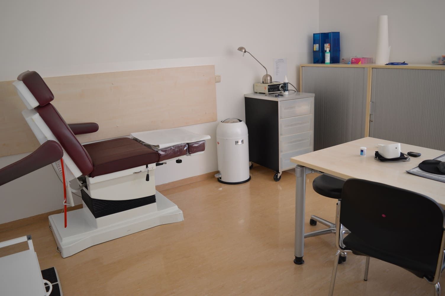 Medizinischer Untersuchungsraum mit Liegestuhl, Schreibtisch und medizinischer Ausrüstung.
