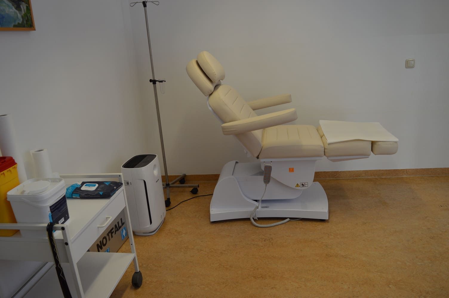 Beiger medizinischer Stuhl in einem Raum, neben einem Wagen, einem Luftreiniger und Geräten.