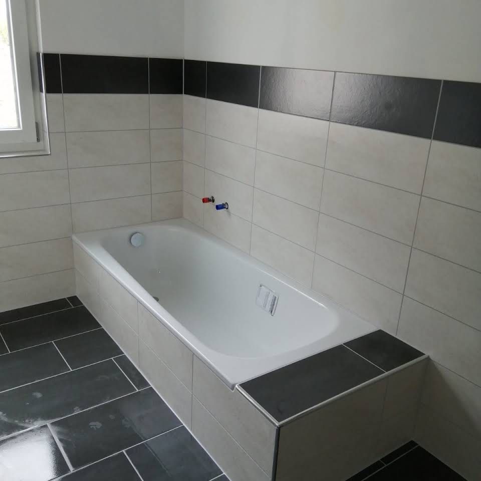 Eine weiße Badewanne in einer Ecke, umgeben von cremefarbenen Wandfliesen, einem schwarzen Fliesenrand und dunkelgrauen Bodenfliesen.