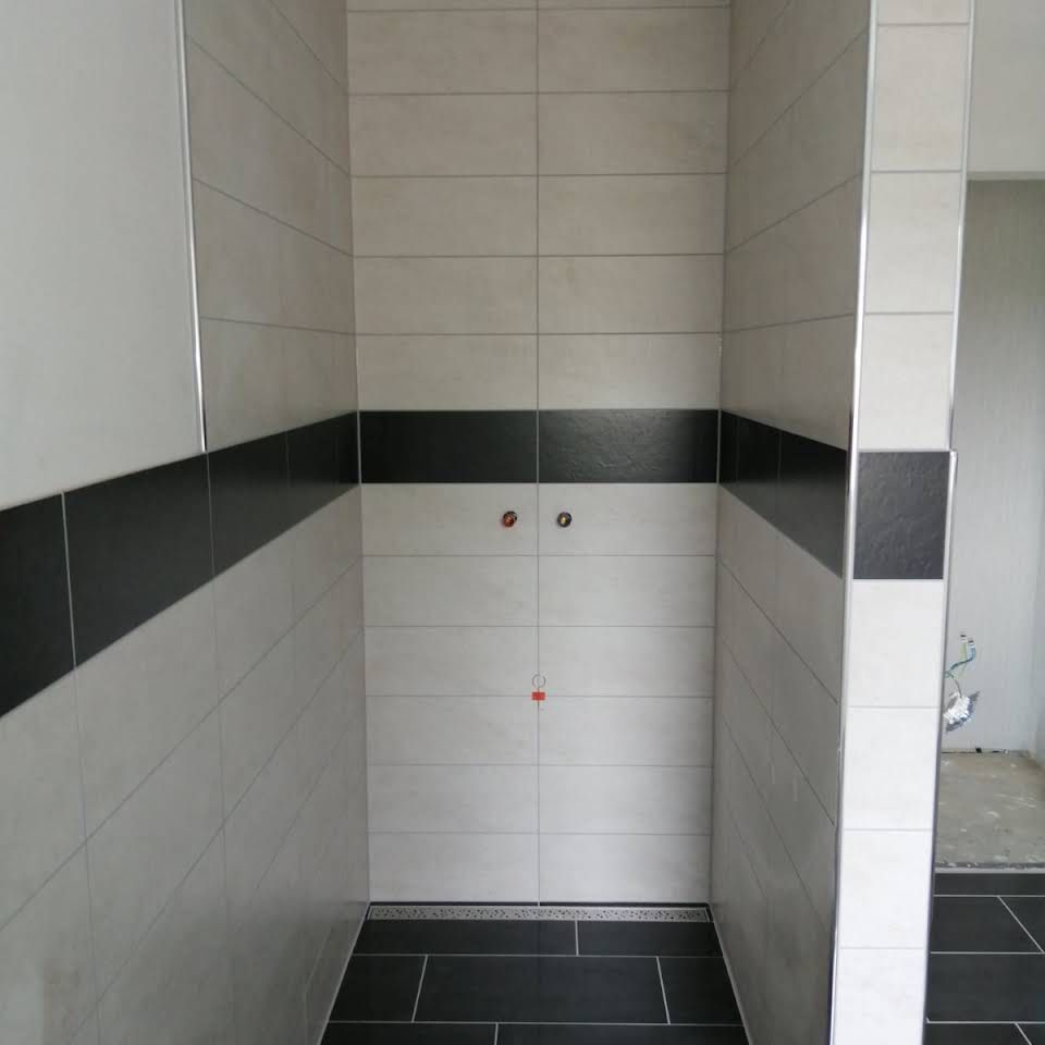 Eine begehbare Dusche mit hellgrauen Wänden, einem horizontalen schwarzen Fliesenstreifen, dunkelgrauen Bodenfliesen und Chromleisten.