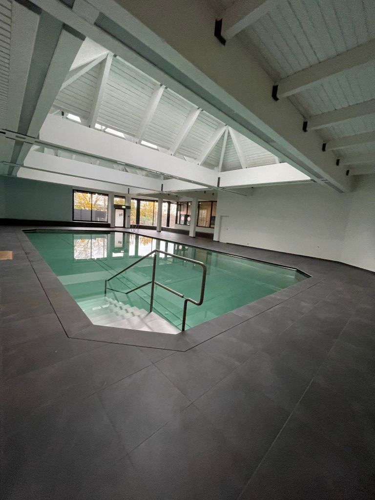 Hallenbad mit gefliestem Beckenrand, grünem Wasser, Edelstahlgeländern und einer schrägen, holzgetäfelten Decke mit Oberlichtern.