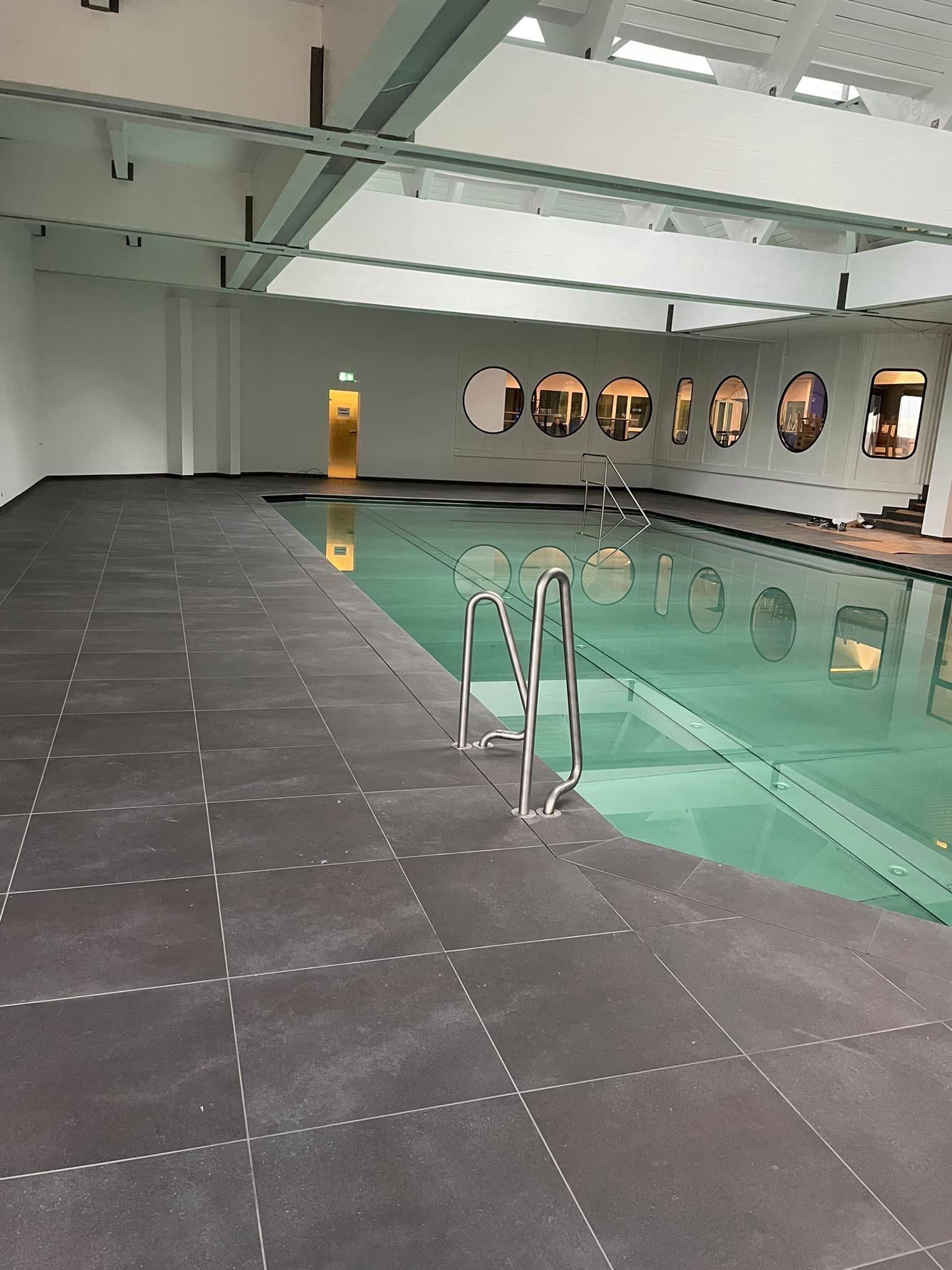 Ein rechteckiges Hallenbad mit grauen Fliesen, einer Metallleiter und runden Spiegeln an der Rückwand.