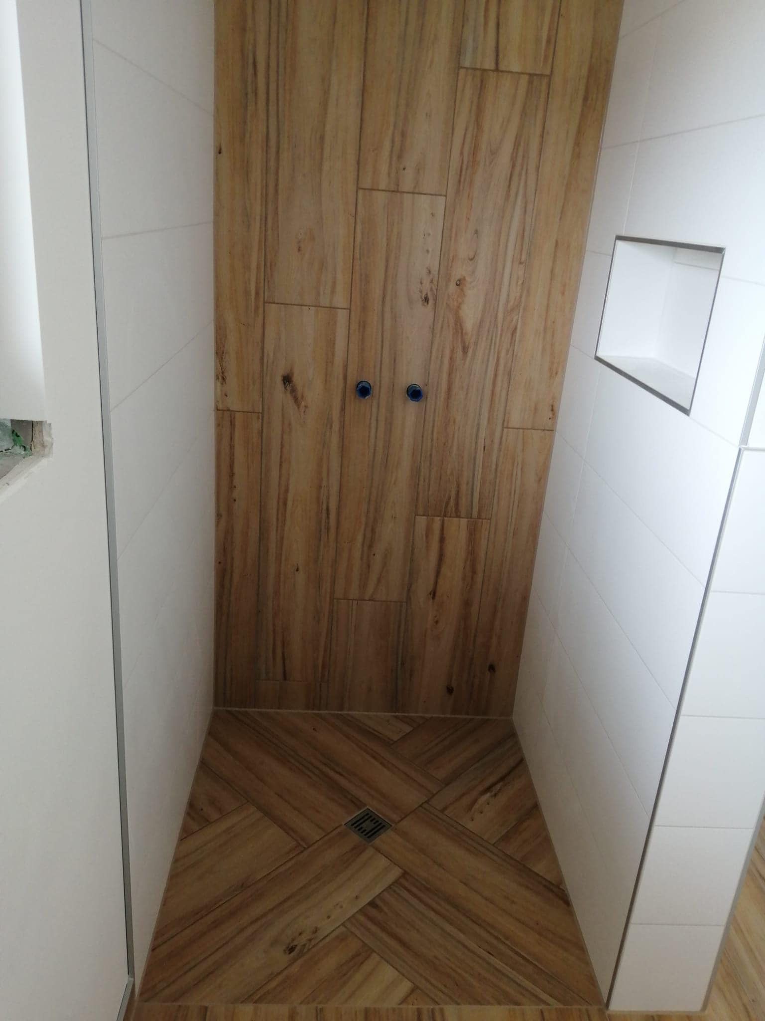 Eine begehbare Dusche mit vertikalen Fliesen in Holzoptik an der Rückwand, Fischgrätfliesen in Holzoptik auf dem Boden und weißen Seitenwänden.