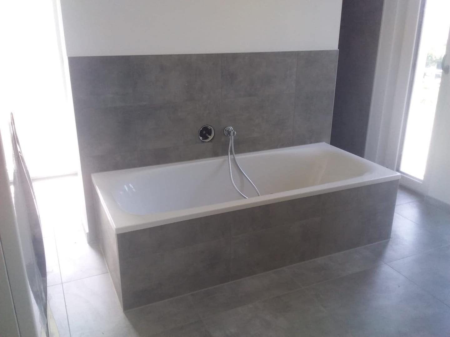 Eine eingebaute weiße Badewanne mit Handbrause vor einer grau gefliesten Wand in einem modernen, in neutralen Tönen gehaltenen Badezimmer.