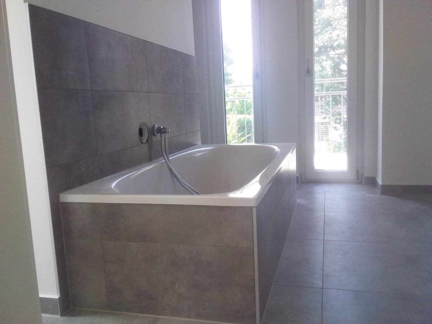 Eine weiße Badewanne vor einer Wand mit dunkelgrauen Steinfliesen in einem hellen Badezimmer neben einer gläsernen Terrassentür.