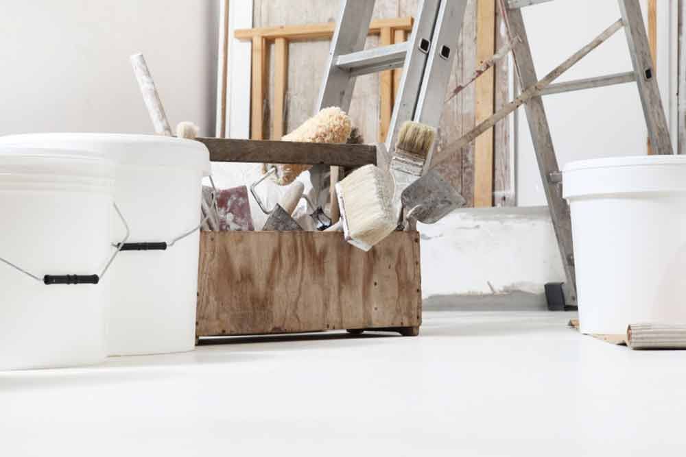 Malutensilien: Farbeimer, ein hölzerner Werkzeugkasten mit Pinseln und eine Leiter auf einem weißen Boden.
