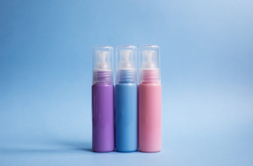 Tres frascos de spray cosmético en tonos pastel —morado, azul y rosa— sobre un fondo azul claro.
