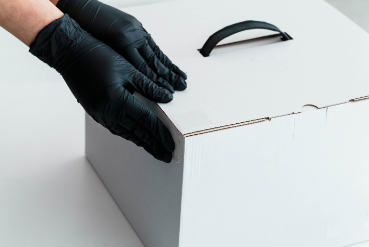 Manos enguantadas de negro abren una caja de cartón blanca con asa negra.
