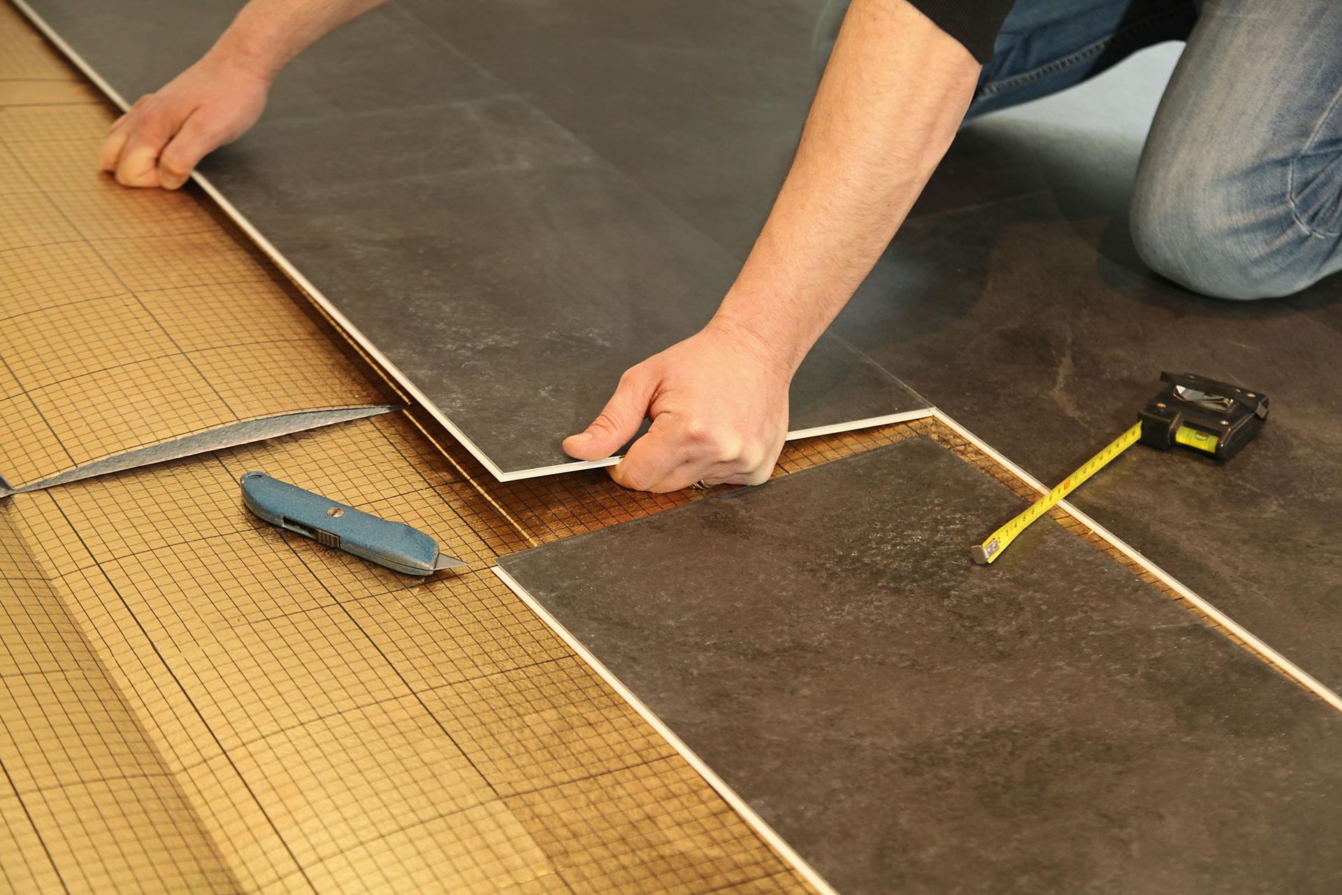 Des mains posent du carrelage foncé sur un sous-plancher, avec des outils de mesure à proximité
