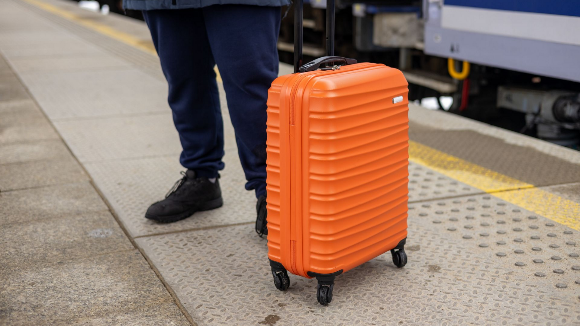 Une personne se tient à côté d'une valise orange sur le quai d'une gare.
