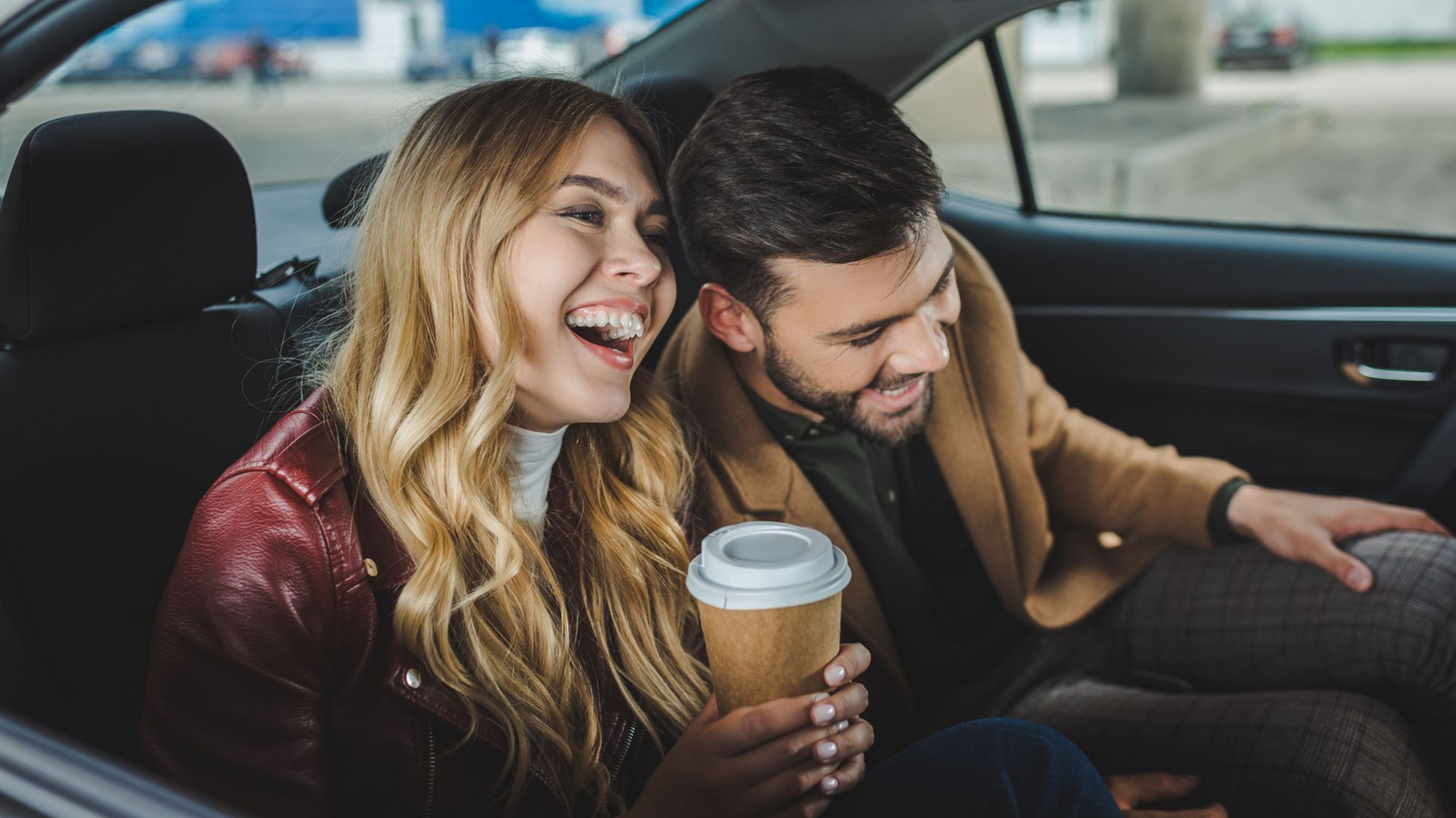 Un couple rit en voiture ; la femme tient une tasse de café. Elle porte une veste rouge, lui un manteau beige.
