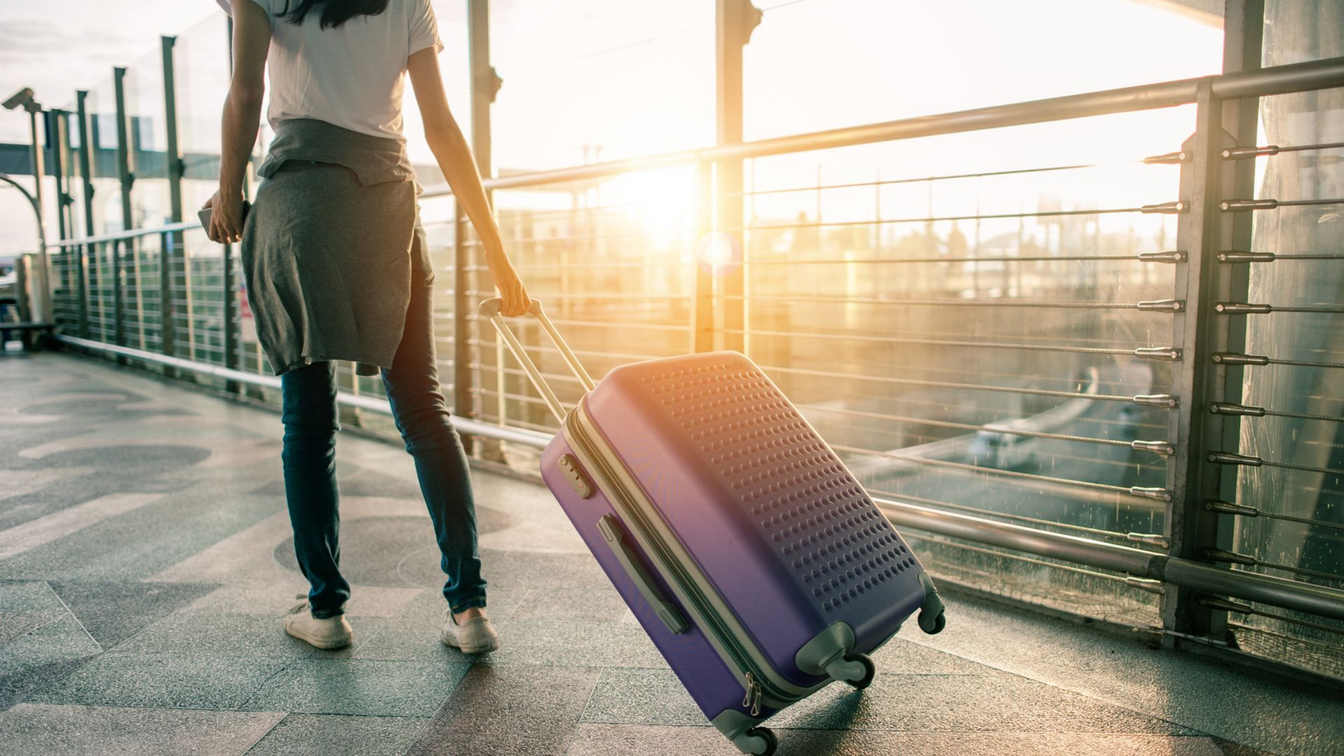 Une personne tirant une valise violette à l'extérieur, en plein soleil, près d'une rambarde métallique.