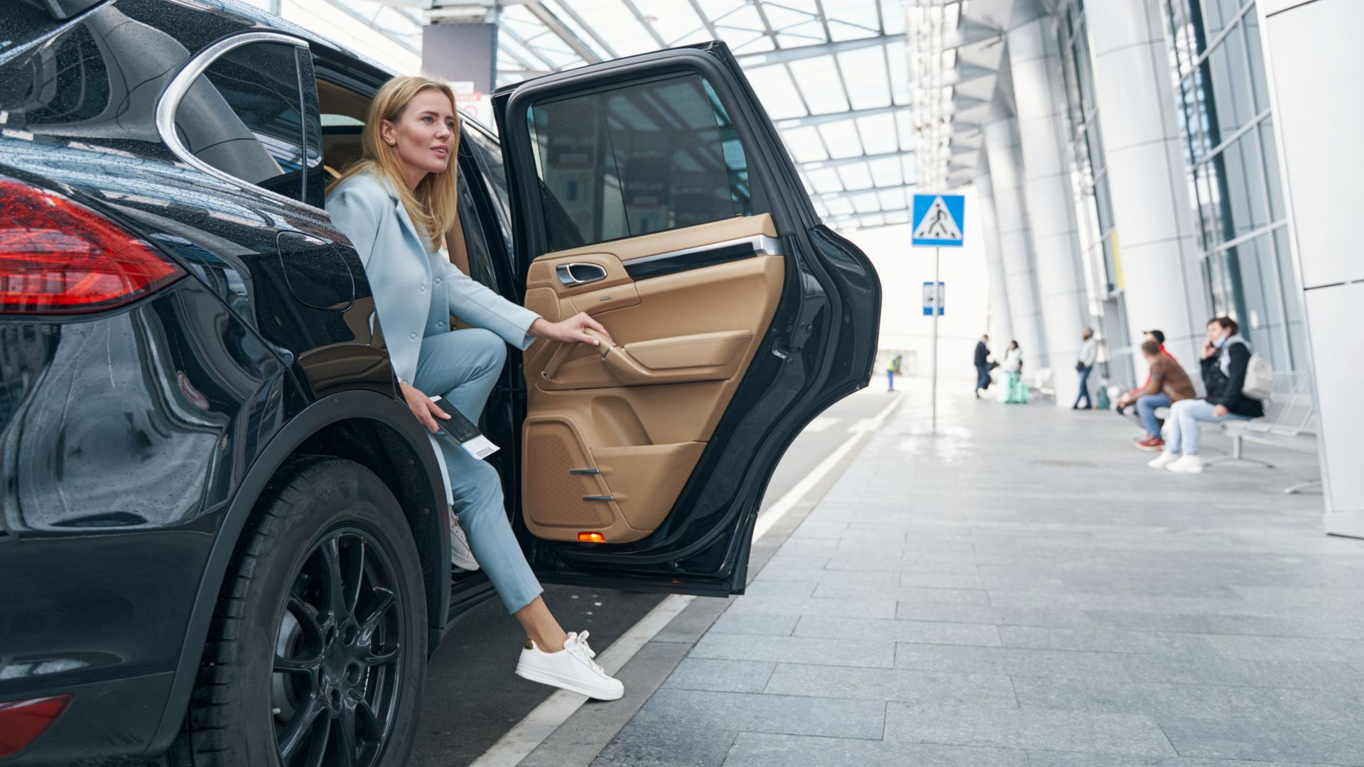 Une femme sort d'un SUV noir à l'aéroport, vêtue d'un jean bleu et d'un blazer.