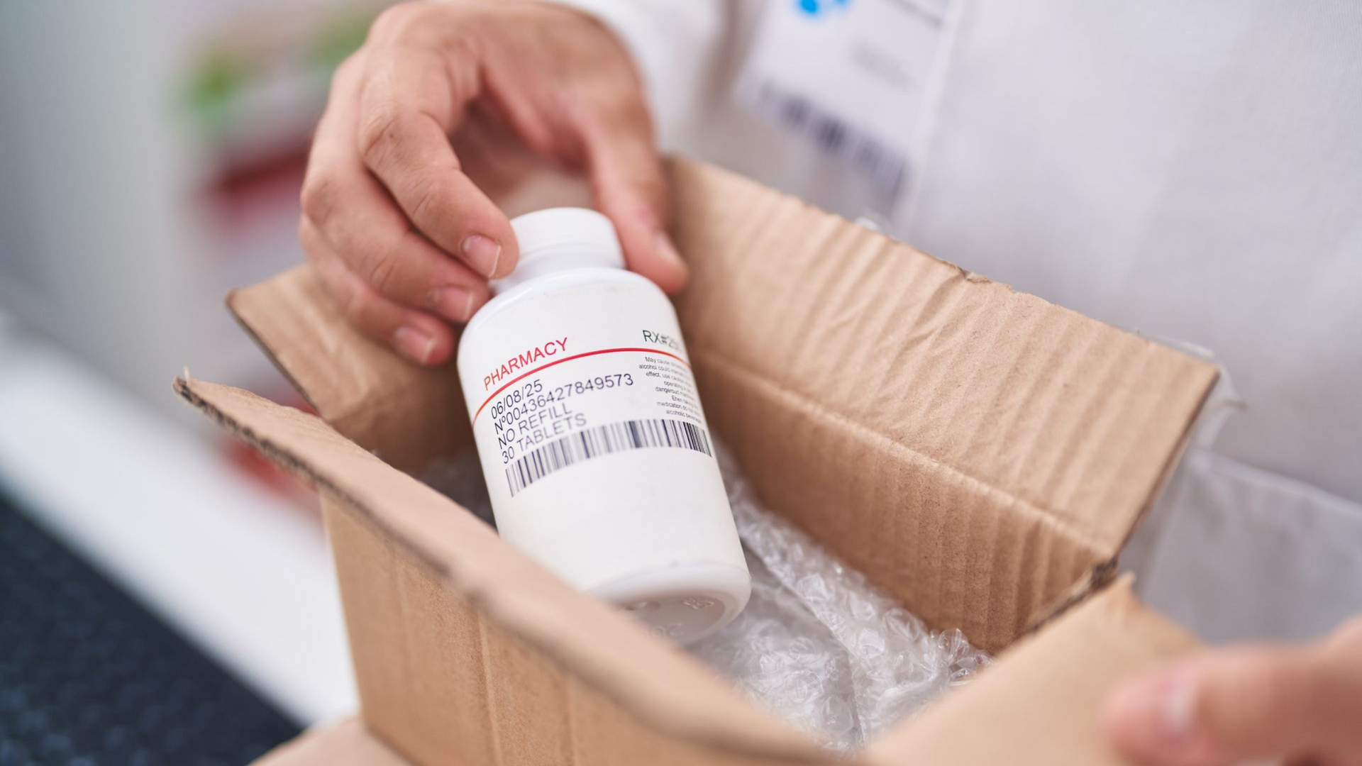 Une personne en blouse blanche ouvre une boîte en carton contenant un flacon de médicament blanc avec une étiquette.