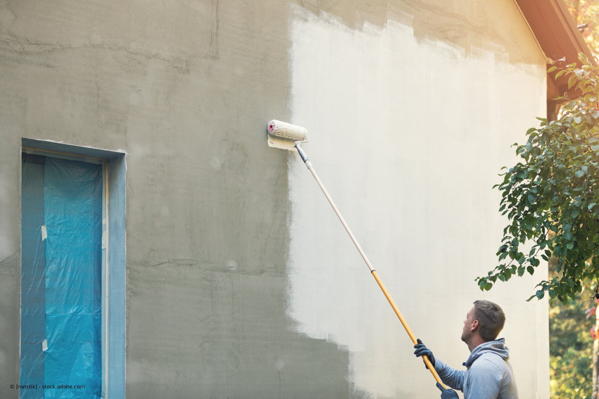 Eine Person streicht eine Außenwand mit einer Farbrolle. Die Wand ist teilweise gestrichen.