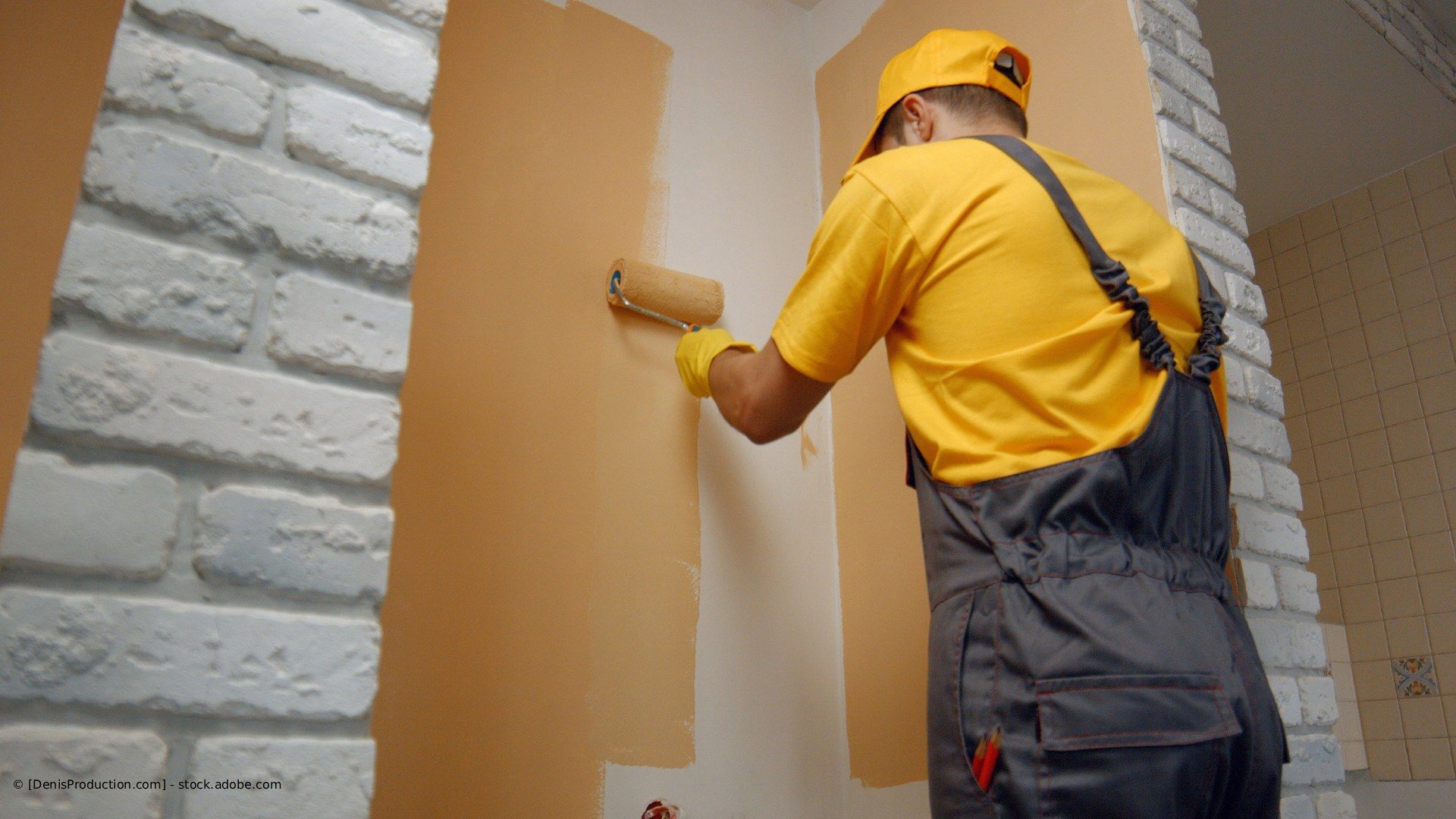 Eine Person in gelbem Hemd und Latzhose streicht mit einer Farbrolle eine Wand.