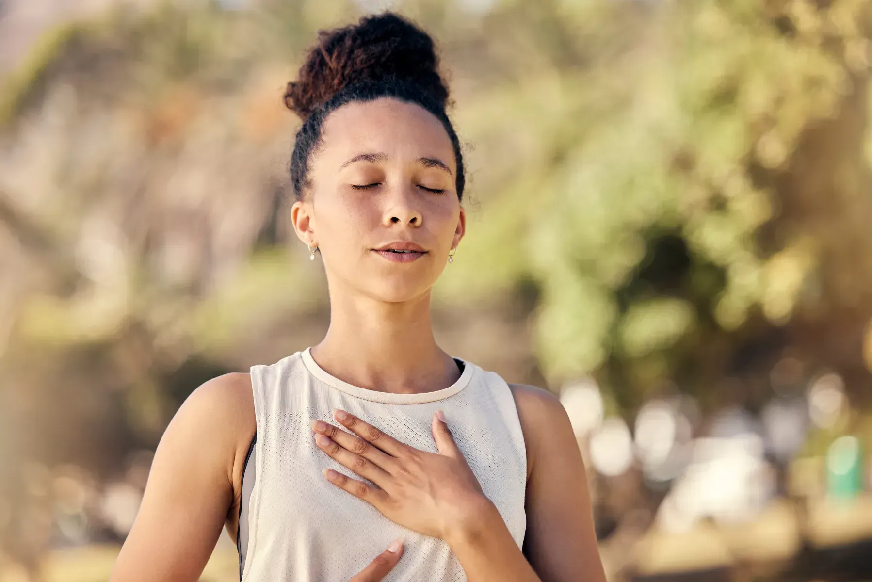 Una mujer está meditando en un parque con los ojos cerrados y las manos en el pecho.