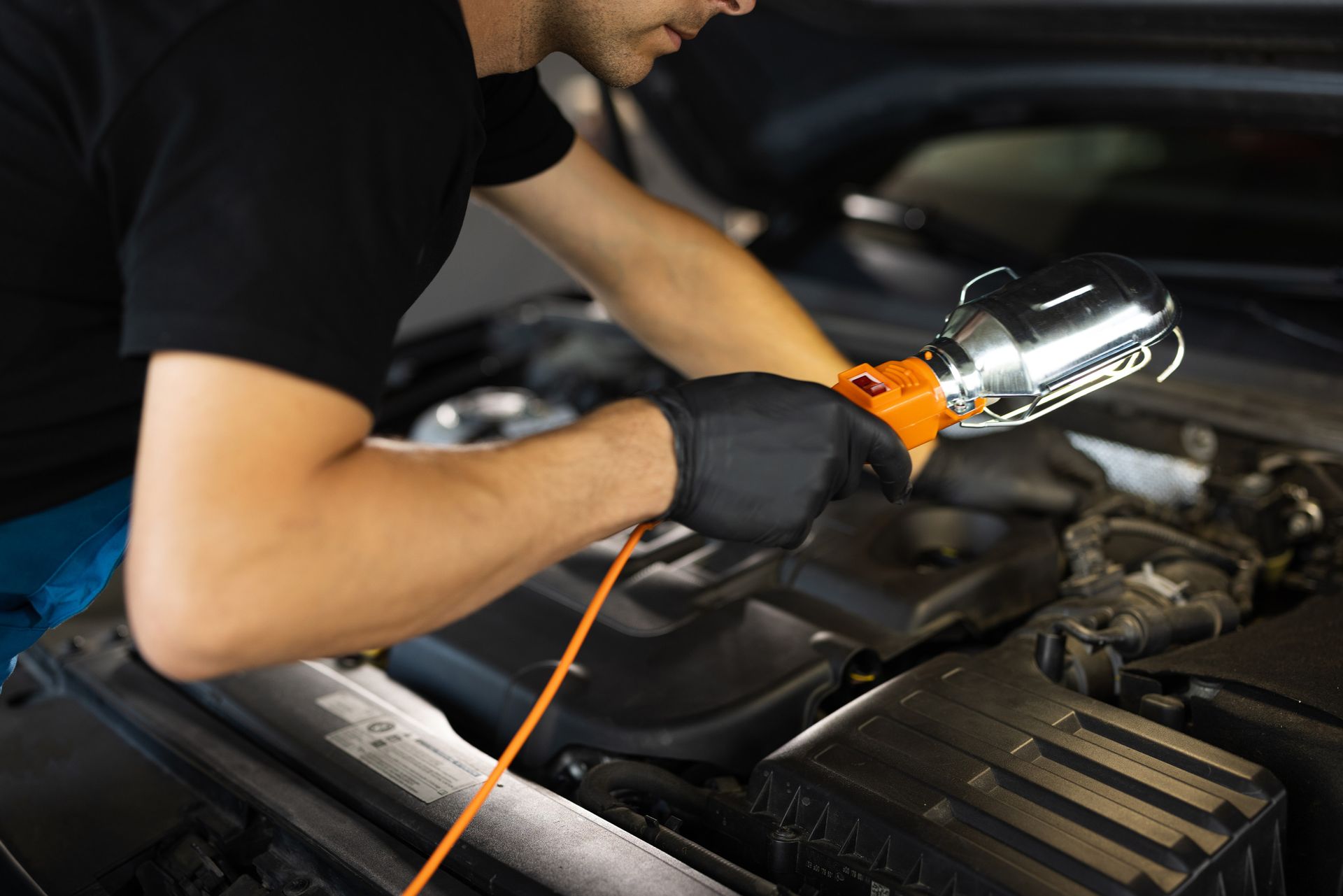 Mecánico inspeccionando el motor de un coche con una luz de trabajo. Lleva guantes y camisa negros en un taller.