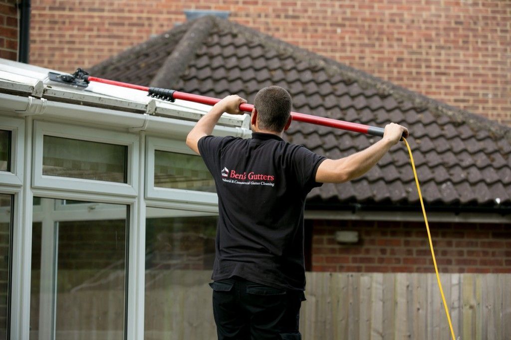 Man on ladder using a long pole to clean a house gutter beside a brick building