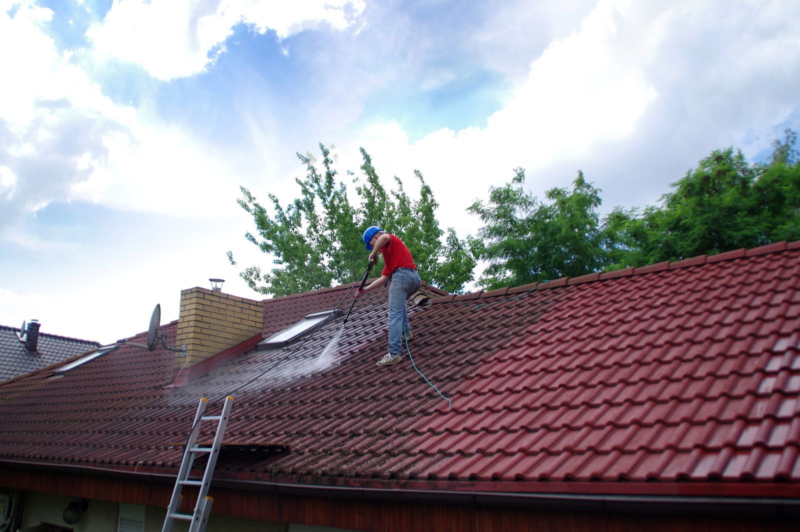 Worker on a red tiled roof repairing shingles beside a ladder under a partly cloudy sky