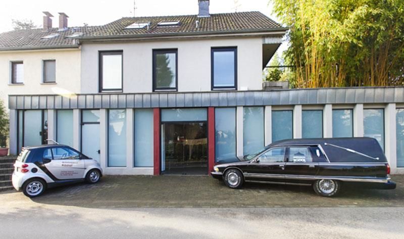 Ein Smart und ein Leichenwagen stehen vor einem Gebäude