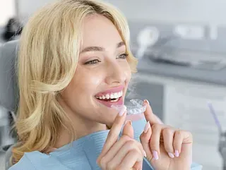 Una mujer está sentada en una silla dental sosteniendo un aparato dental transparente en su boca.